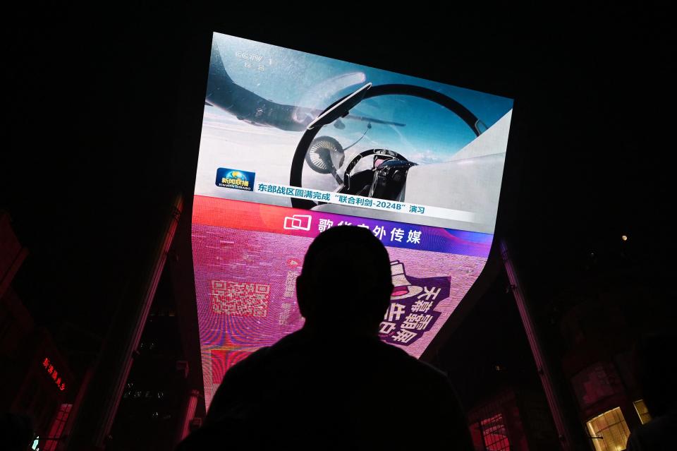 A man watches a big screen showing a pilot fly a fighter jet above a cloudy and blue sky.