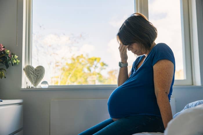 Pregnant woman sits on a bed, holding her forehead in contemplation by a window