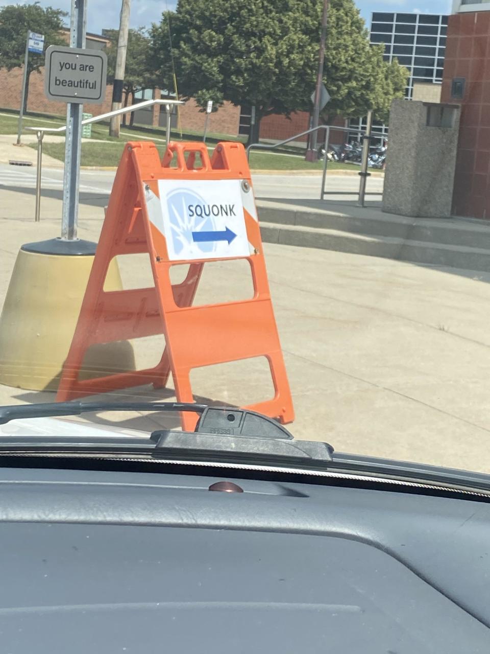 Orange traffic sign on a sidewalk with "SQUONK" and an arrow. A small sign nearby reads "you are beautiful."