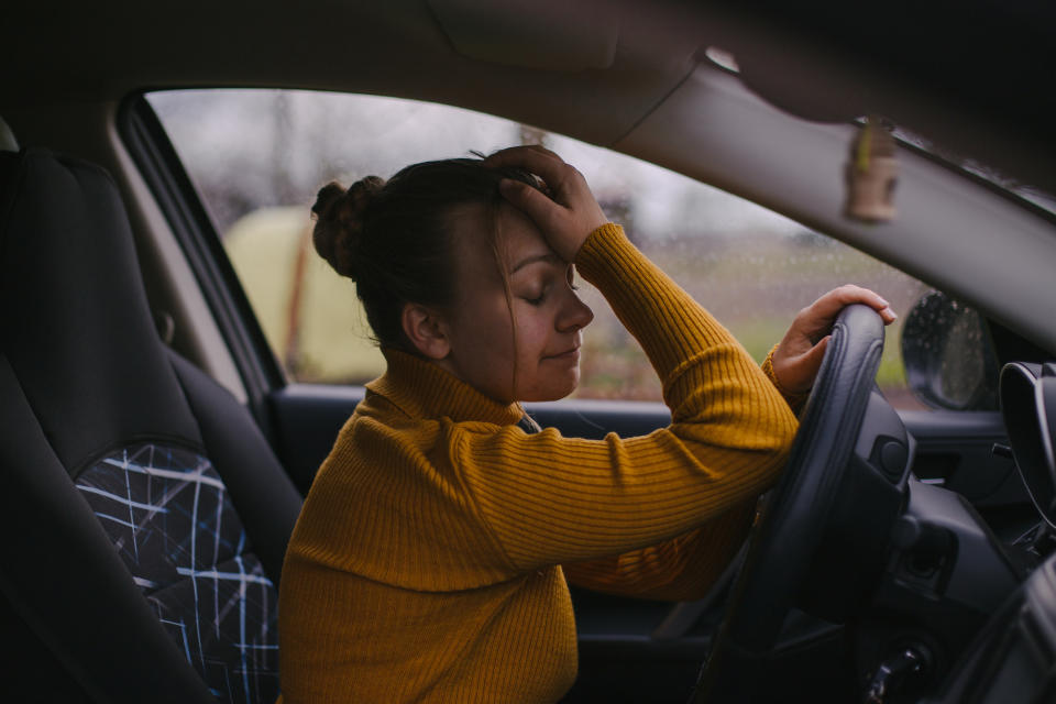 A person with closed eyes rests their head on a steering wheel, looking contemplative inside a car