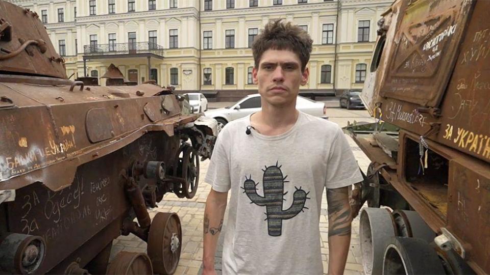 Oleksii standing between two military vehicles in Kyiv after his rescue. He has short brown hair and is wearing a white T-shirt with a picture of a cactus on it.