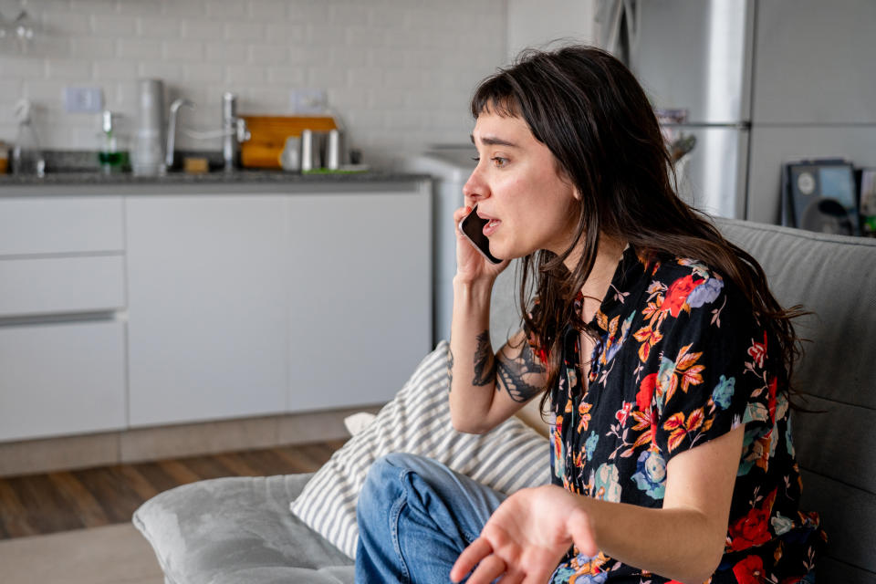 A woman in floral top sits on a couch, talking on the phone with a concerned expression in a modern kitchen/living room space