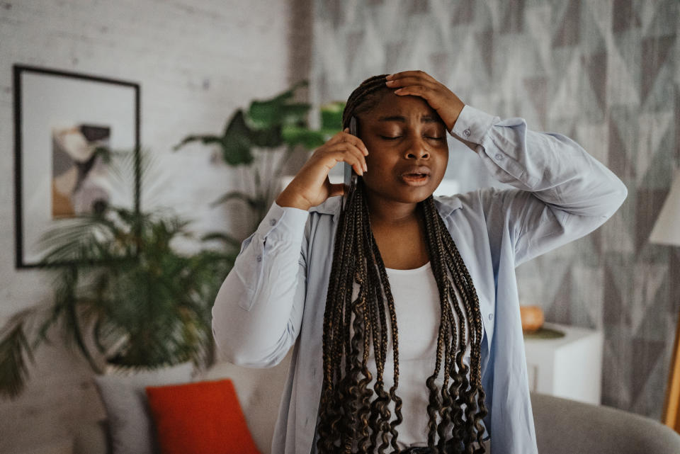 Person with long braids, holding phone to ear, eyes closed, hand on forehead, appears stressed, standing in a living room with plants and framed art