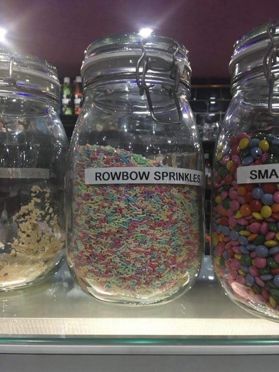 Jars on a shelf with labels: "Rowbow Sprinkles" filled with multicolored sprinkles, others with small multicolored candies