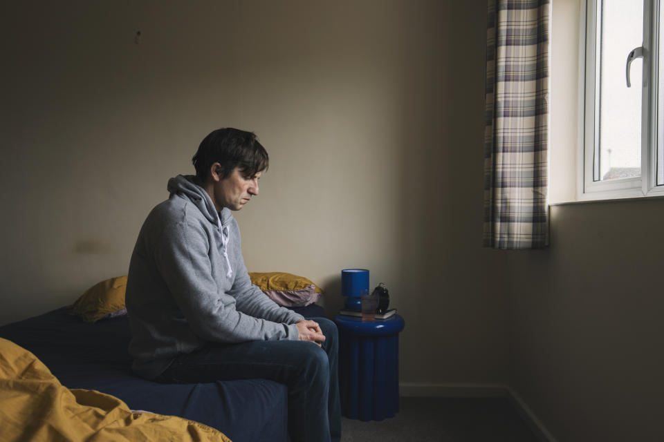 A person sits on a bed in a dimly lit room, appearing pensive and alone, beside a window and a small table with items on it