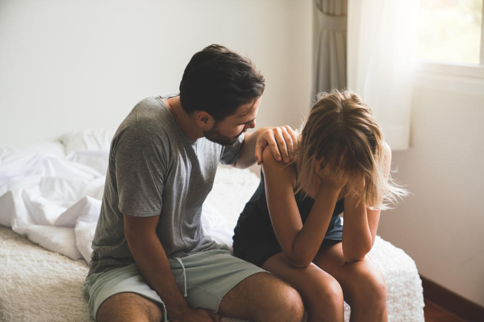 A man consoles a distressed woman sitting on a bed, showing emotional support in a bedroom setting