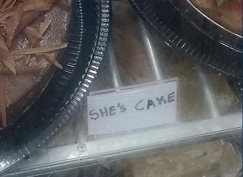 A bakery display with a handwritten sign reading "SHE'S CAKE" next to a cake