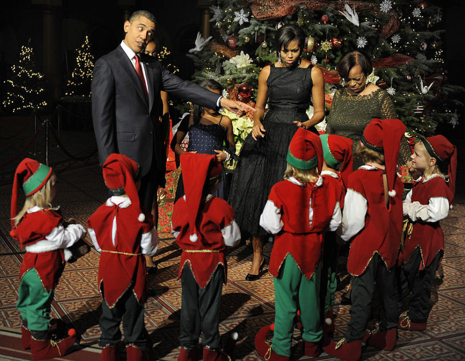 A man in a suit and a woman in a formal black dress interact with children dressed as elves in front of a decorated Christmas tree