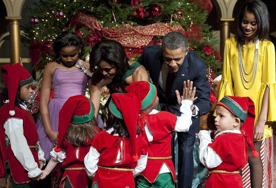 A family greeting children dressed as elves next to a Christmas tree. A woman in a formal dress and a man in a suit are interacting with the children