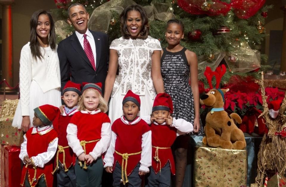 A group photo with adults including a man in a suit and a woman in a lace dress, posing with children in elf costumes and a large stuffed dog near a Christmas tree