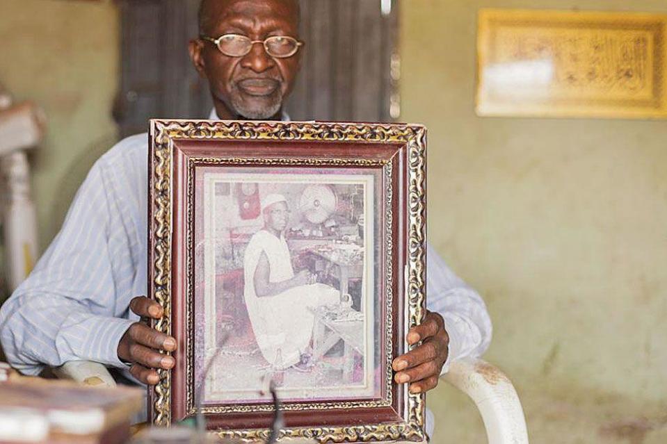 Bala Muhammad, wearing wire-rimmed glasses and a long-sleeved pin-stripped blue shirt, sits in a white-painted wooden chair holding up a wide-wooden framed black-and-white photograph of his father, who is pictured looking at the camera as he sits at his workshop bench. He is wearing a sleeveless boubou. A desk fan can be seen in the background and a pendulum clock hangs on the wall