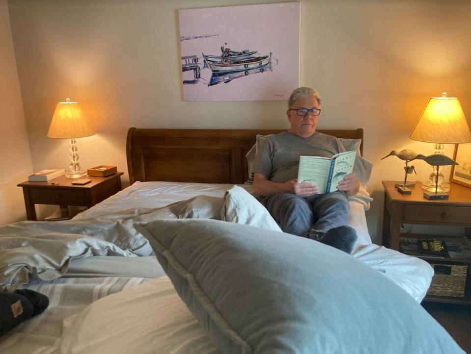 Man sitting on a bed, wearing glasses, reading a book under soft lighting. A painting of boats hangs above the bed