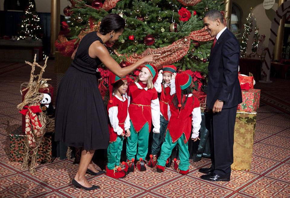 Two adults in elegant clothing interact warmly with children dressed as elves in front of a festive Christmas tree