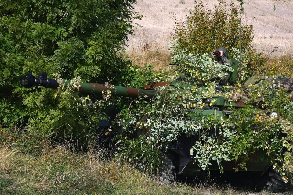 A soldier looks through binoculars from the turret of an AMX-10RC wheeled tank destroyer concealed in the foliage.