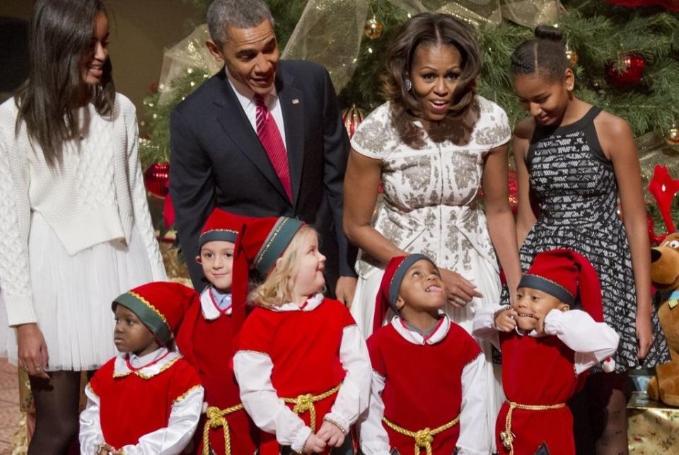 Group of adults and children at a festive event. Adults in formal attire, children in elf costumes. Holiday decorations in the background