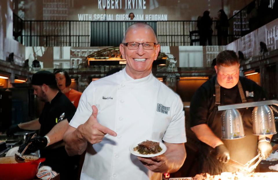 Chef in a kitchen holds a plate of food, smiling, while two chefs work in the background on meal preparations