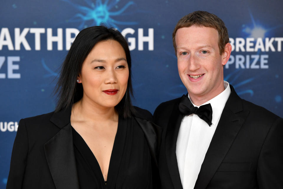 Two people smile on a blue-themed red carpet. The woman wears a black blazer and dress, and the man wears a black tuxedo and bow tie