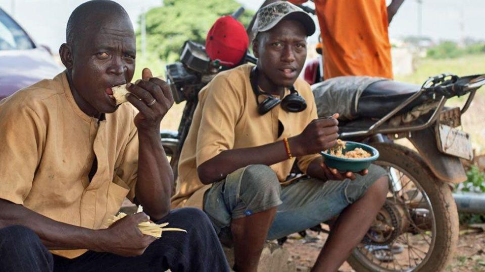 Two graveyard workers sitting down under a tree eating food at lunchtime - behind them a motorbike can be seen