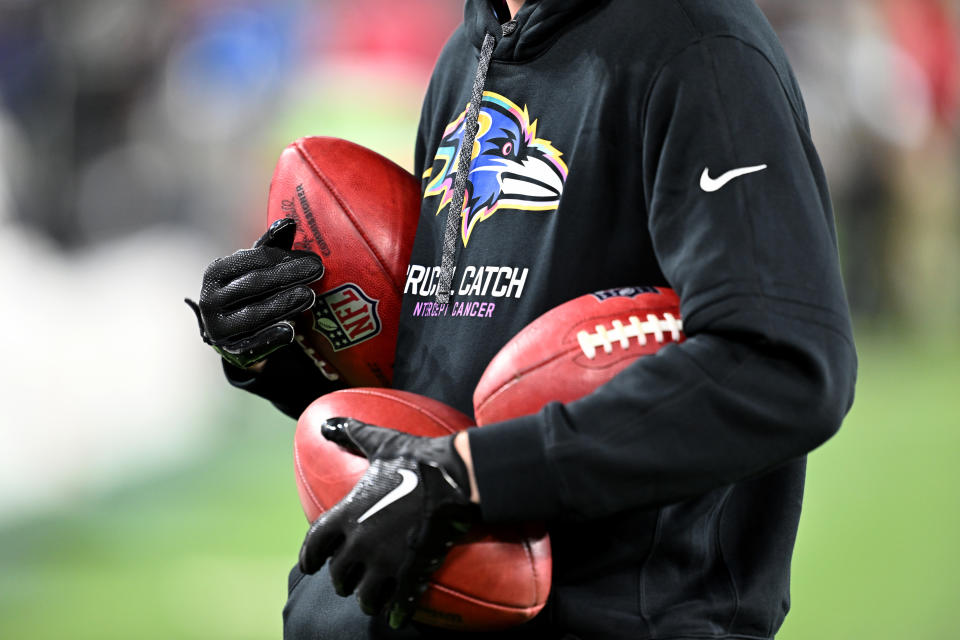 Person holding multiple footballs wearing a hoodie with a team logo and gloves at a sports event