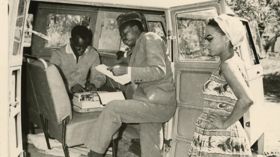 Pierre Mulele, one foot inside a van, peruses a piece of paper. A man inside the van uses a typewriter, while Andrée Blouin stands outside the van with her hands on her hips.