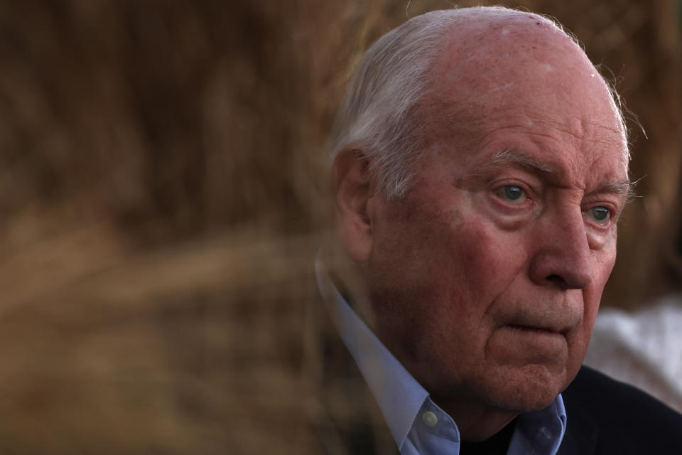 Elderly man with thoughtful expression, wearing a dark jacket over a collared shirt. Background suggests an outdoor setting