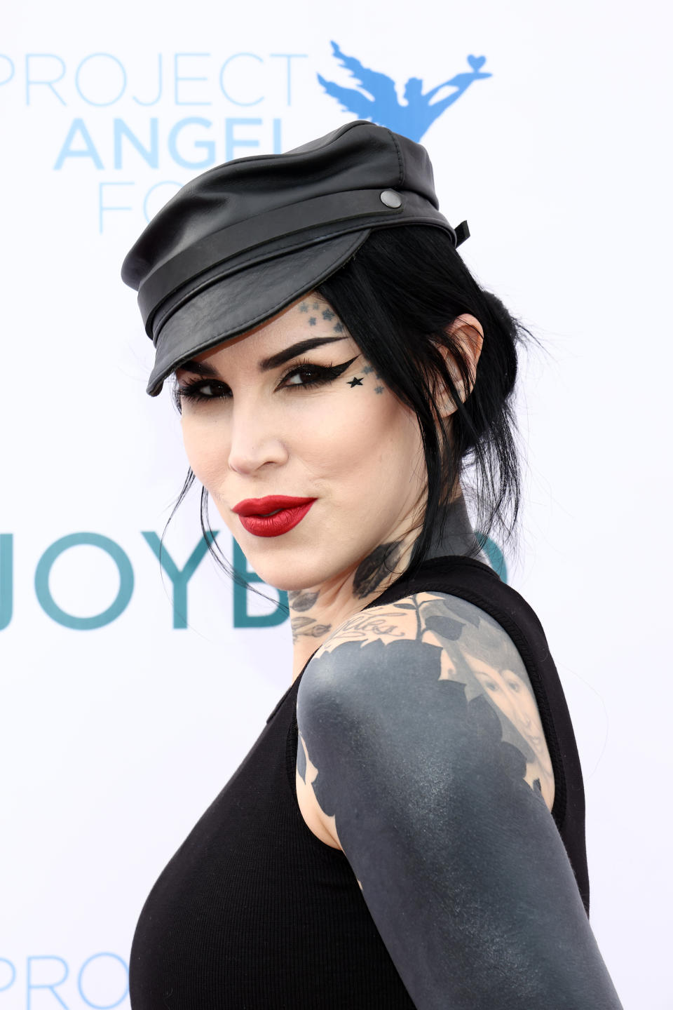 Person with tattoos and black hat poses confidently at an event, wearing a sleeveless top with bold makeup and star details near the eye