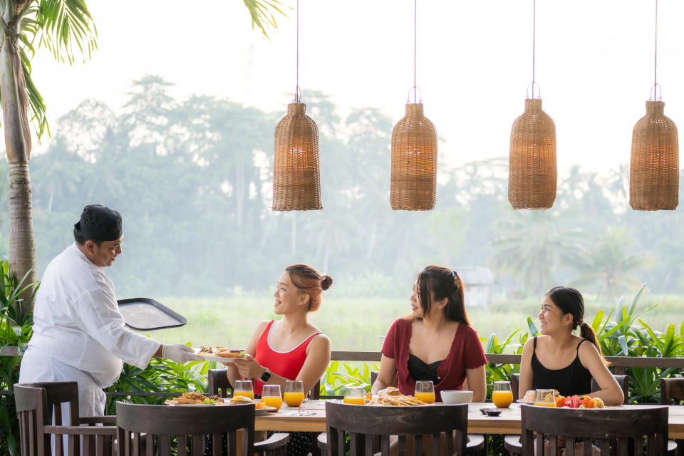 Three women enjoy a private breakfast on a patio while a server in a chef hat delivers food