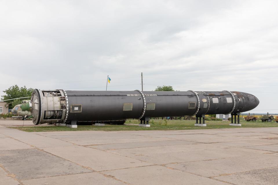 A large grey missile is seen on its side resting on supports above a tarmac and grass ground and a grey sky behind it