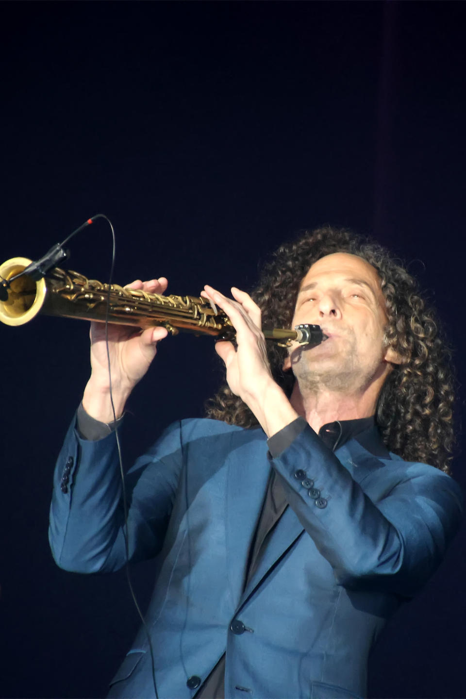 Person playing a saxophone on stage, wearing a suit, with curly hair