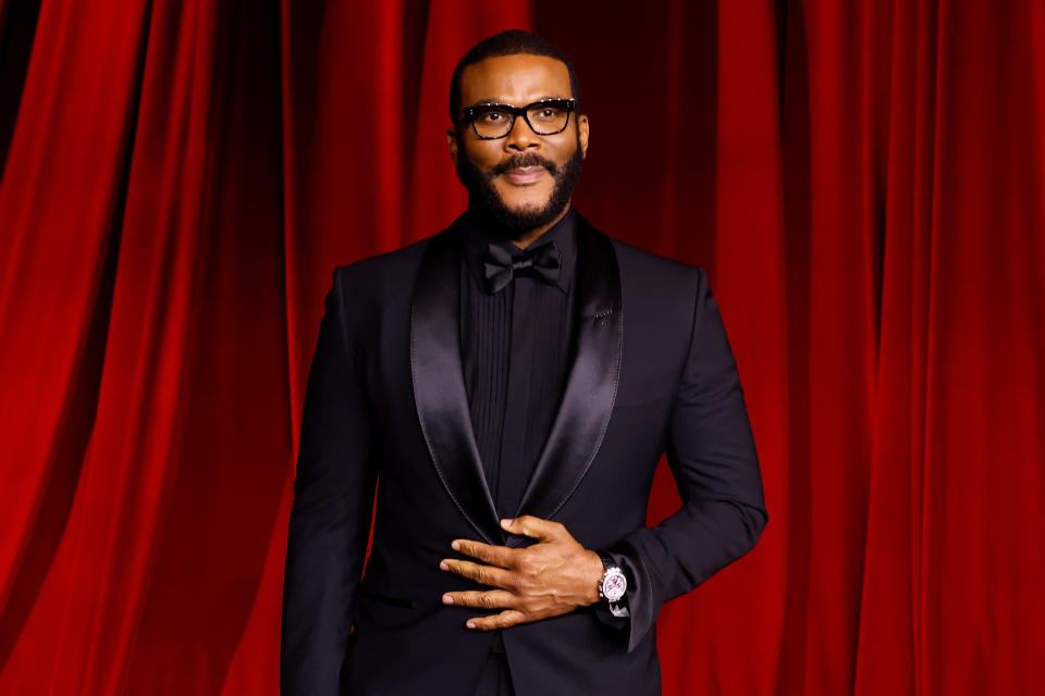 Person in a sleek black tuxedo with black tie, standing in front of a curtain backdrop