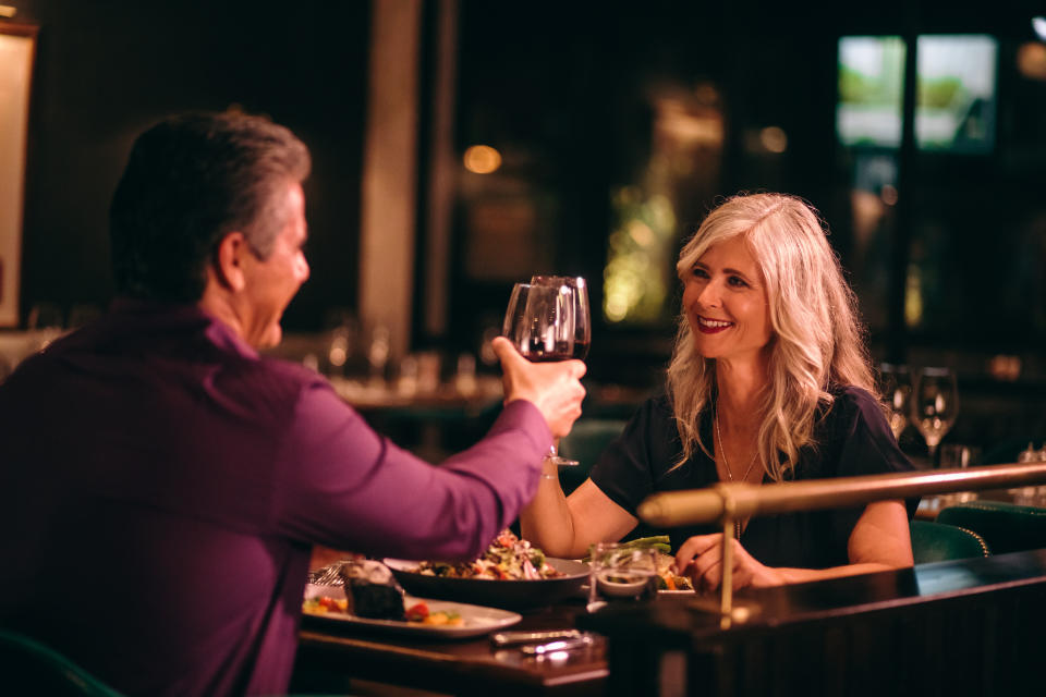 A couple sits at a restaurant table, smiling and toasting with wine glasses