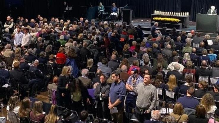 A seated crowd in a large venue. Some members of the crowd are stood with their backs to the stage. One member of the crowd is being grabbed by a woman. 