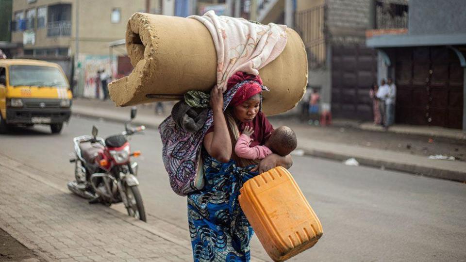 A woman wearing a printed blue wrap walks, slightly hunched forward as she holds a baby, a yellow jerry can and a rolled foam mattress on her head