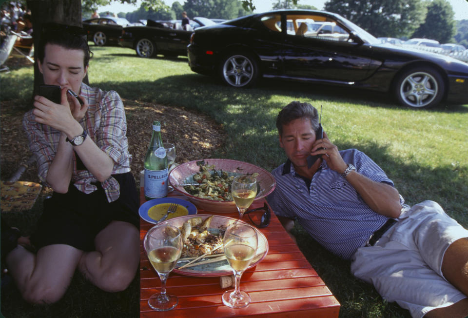 Two people sit at a picnic with fancy-looking food and drinks while one talks on his cell phone