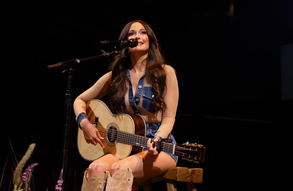 Performer singing on stage, playing an acoustic guitar, wearing a casual denim outfit and boots. Stage light shines on them