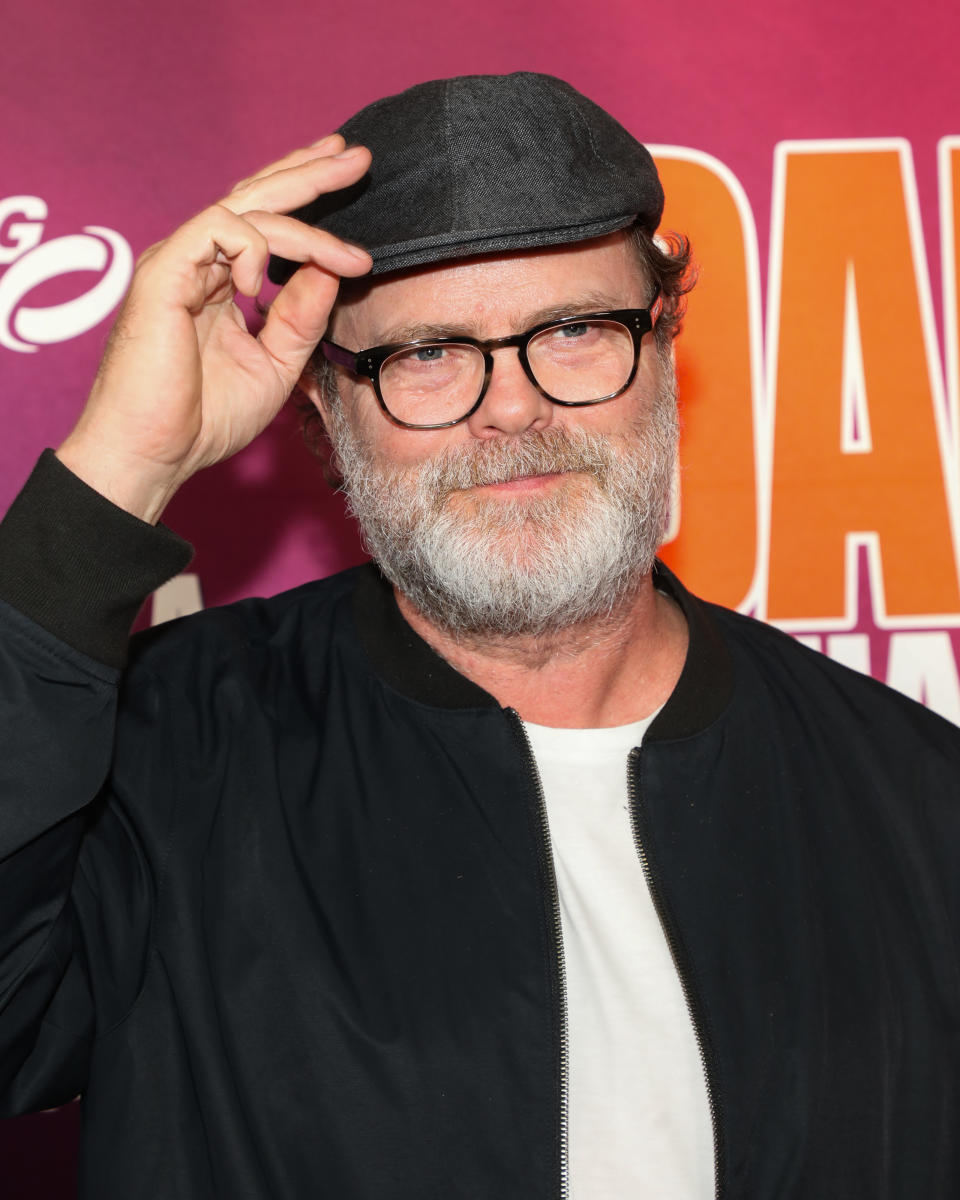 Person with glasses, beard, and a cap on a step-and-repeat background. Wears a casual jacket over a T-shirt, holding the cap's brim