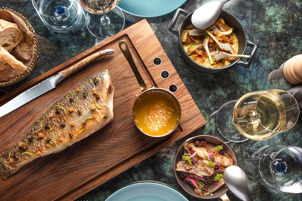 A set marble table with fried whole fish on a wooden cutting board, sauce in a pan on the side and vegetable dishes.