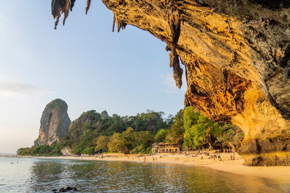 A view from under a cliff arch onto a busy beach at sunset with hills and a forest in the distance.