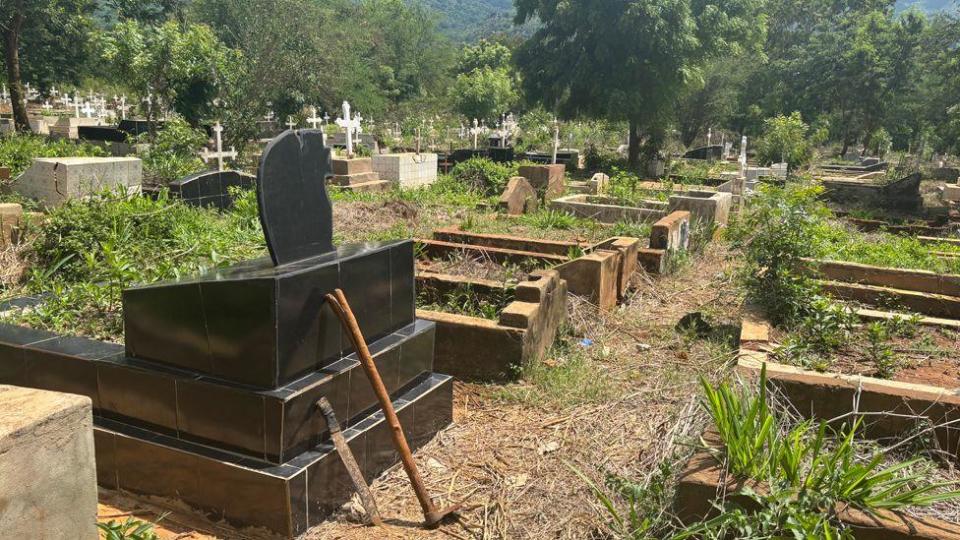 A wide view of a cemetery showing a series of vandalised graves. The cemetery is in a lush setting with green trees and shrubs visible.