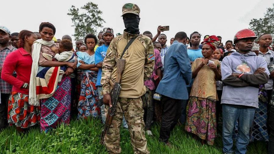 A member of the M23 in a balaclava and with an automatic weapons stands in front of a crowd of civilians.