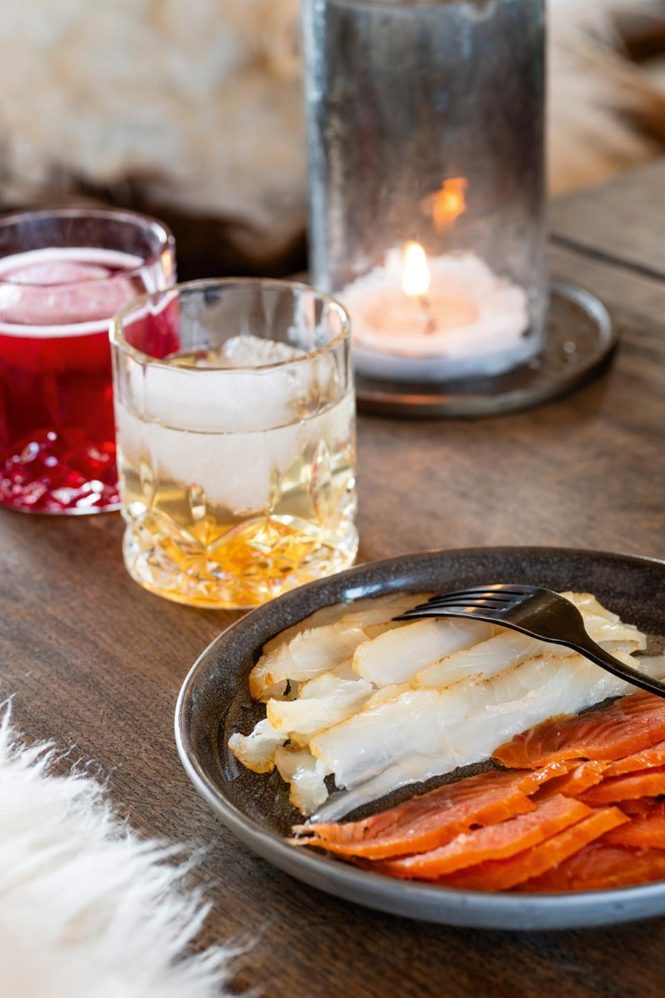 A plate of cured fish with a side of cocktails on ice and a candle in the back.