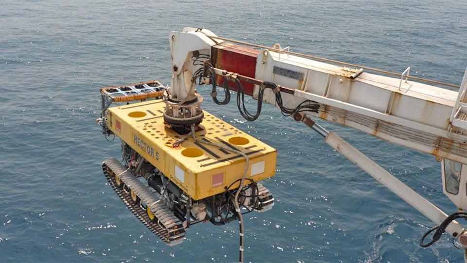 A bulldozer-like yellow remotely operated vehicle (ROV), with words Hector 5 on it, hangs off a crane over the sea.