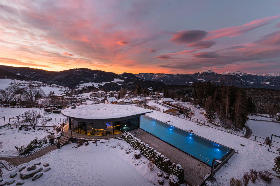 A breathtaking sunset over the snow-covered landscape of Chalet Mirabell, with an illuminated heated infinity pool overlooking the Dolomites.