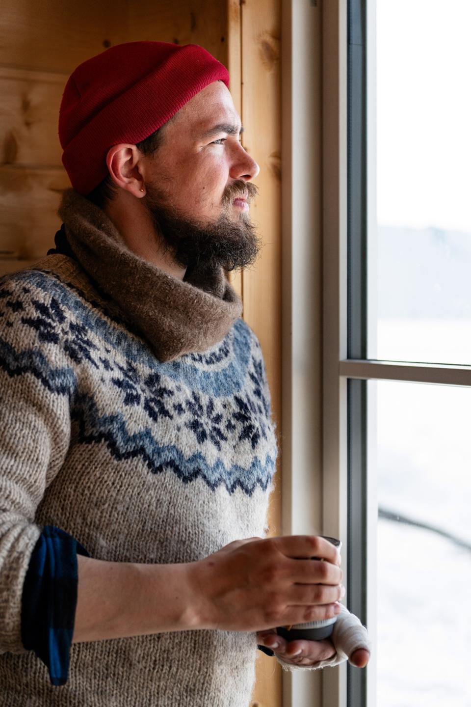 A man in a thick cable-knit jumper and wool hat, holding a hot cup of tea and looking out the window.