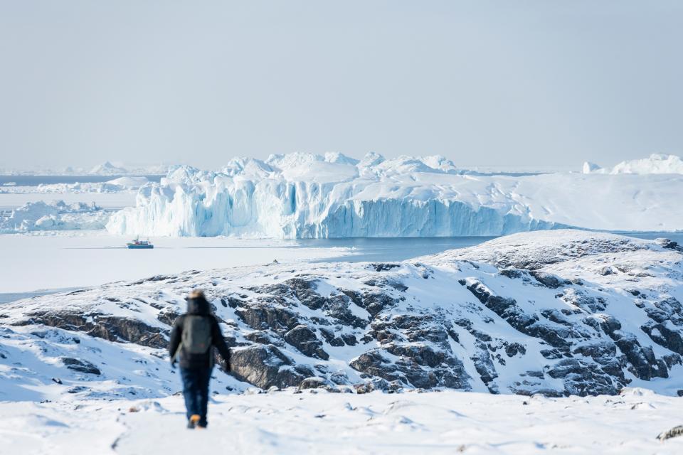 A wide landscape shot looking out at an icy fjord with an imposing iceberg swimming in the sea and a person walking towards it.