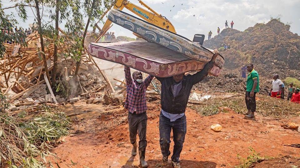 A man holds up his hands to help another man carrying tow mattresses away from Kiteezi waste site after its collapse in August 2024 - Kampala, Uganda