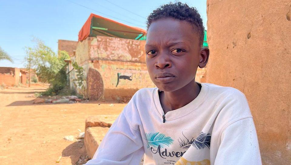 Muzami, the 18-year-old son of Intisar Adam Suleiman, looks directly into the camera lens with a pained expression. He is wearing a white sweat shirt and sitting outside an orange-plastered building on a street in the Haj Yusuf district of Sudan's capital Khartoum - March 2025