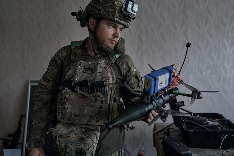 A drone pilotss wearing camouflage gear and a helmet stands holding a door in front of a wall with pale wallpaper