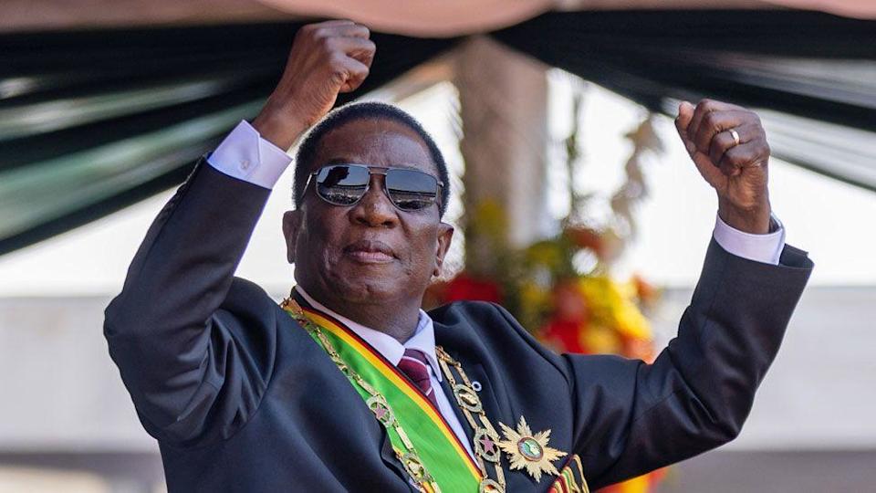 Zimbabwe's President Emmerson Mnangagwa in sunglasses and wearing a suit and sash in the colours of Zimbabwe and a chain and star of office holds up his fists.