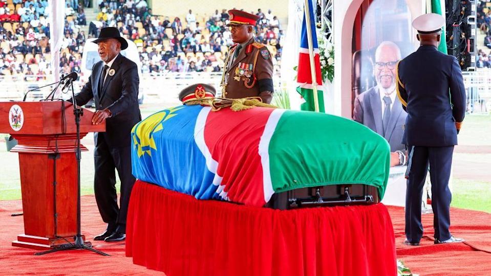 A coffin draped in the Namibian flag stands on a stage as a man in a black suit speaks at a lectern.